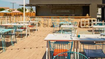 Een terras met tafels en stoelen, een podium met de aanduiding 'LE CABANON', en uitzicht op de omgeving.