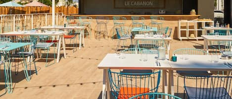 Un área de comedor al aire libre con mesas y sillas, un escenario con el letrero 'LE CABANON', y vistas del entorno.
