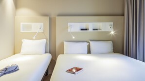 Une chambre d’hôtel avec deux lits, une table de chevet avec une lampe, une étagère fixée au mur avec des verres et un sac, et un rideau à la fenêtre.