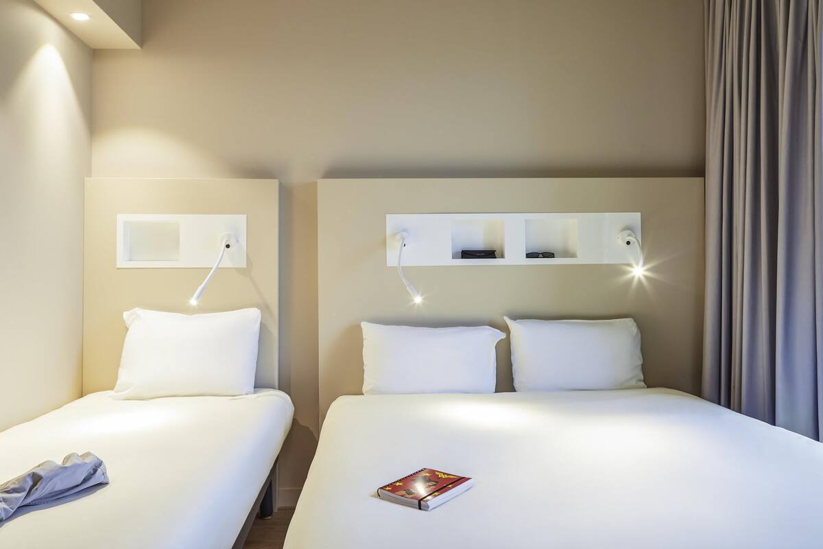 Ein Hotelzimmer mit zwei Betten, einem Nachttisch mit Lampe, einem an der Wand befestigten Regal mit Gläsern und einer Tasche sowie einem Vorhang am Fenster.