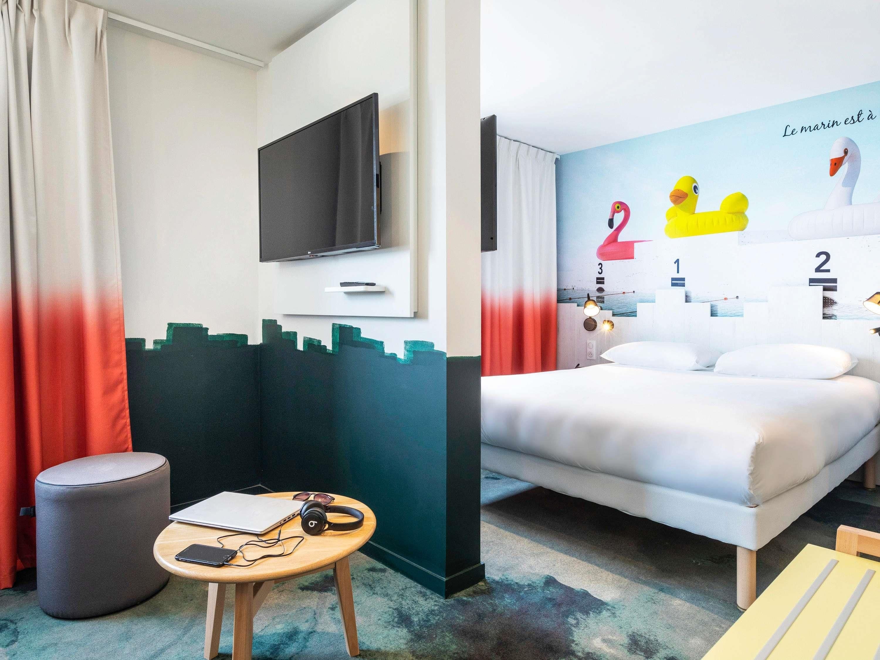 A hotel room with a bed, a TV, a desk with a laptop, headphones, and a mobile phone.