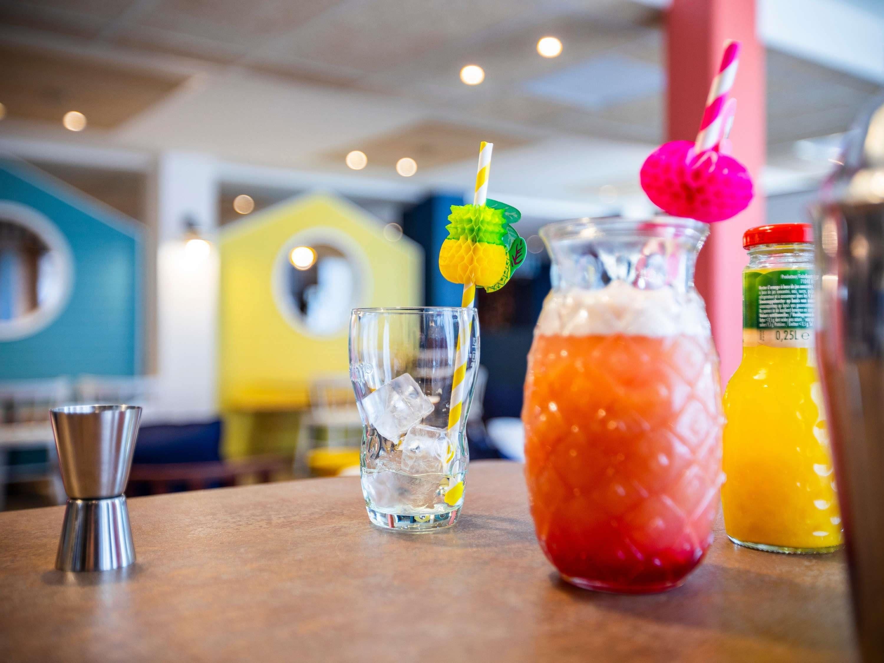 Een baromgeving met een glas ijsthee, een karaf met een rood drankje en een fles sinaasappelsap.
