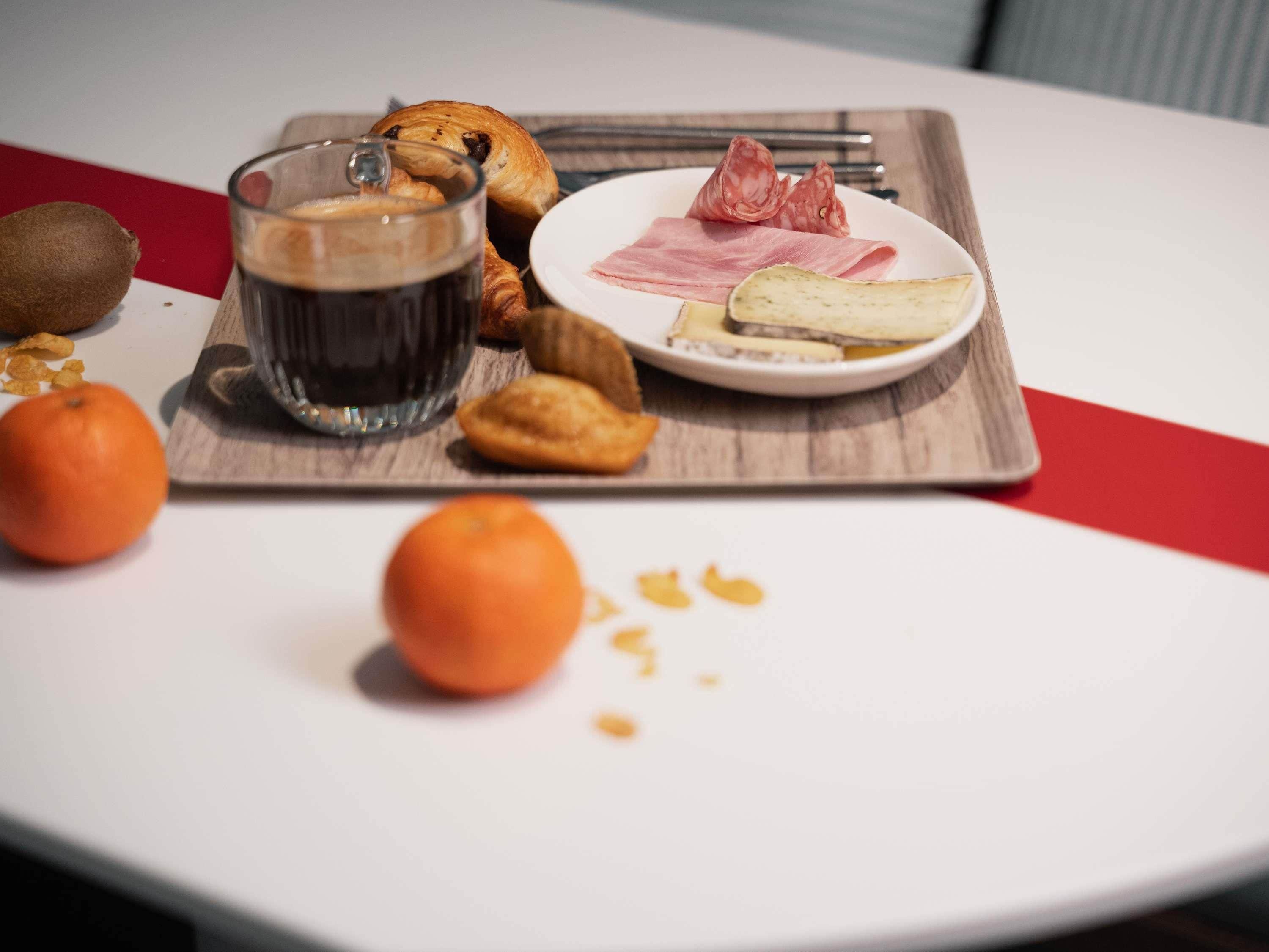 A tray with food items including pastries, cheese, and cured meats, a glass of dark liquid, and two oranges on a white table.