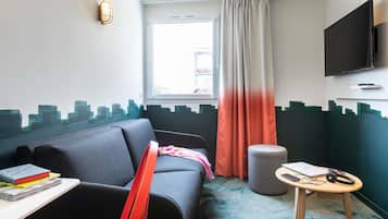 Quarto de hotel compacto com sofá, uma pequena mesa com livros, um banquinho redondo, televisão fixada na parede e uma janela com cortinas.
