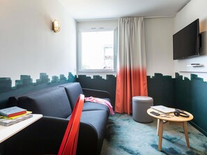A compact hotel room with a sofa, a small table with books, a round stool, a television mounted on the wall, and a window with curtains. - ibis Styles Thonon Les Bains Hotel (Thonon-les-Bains)