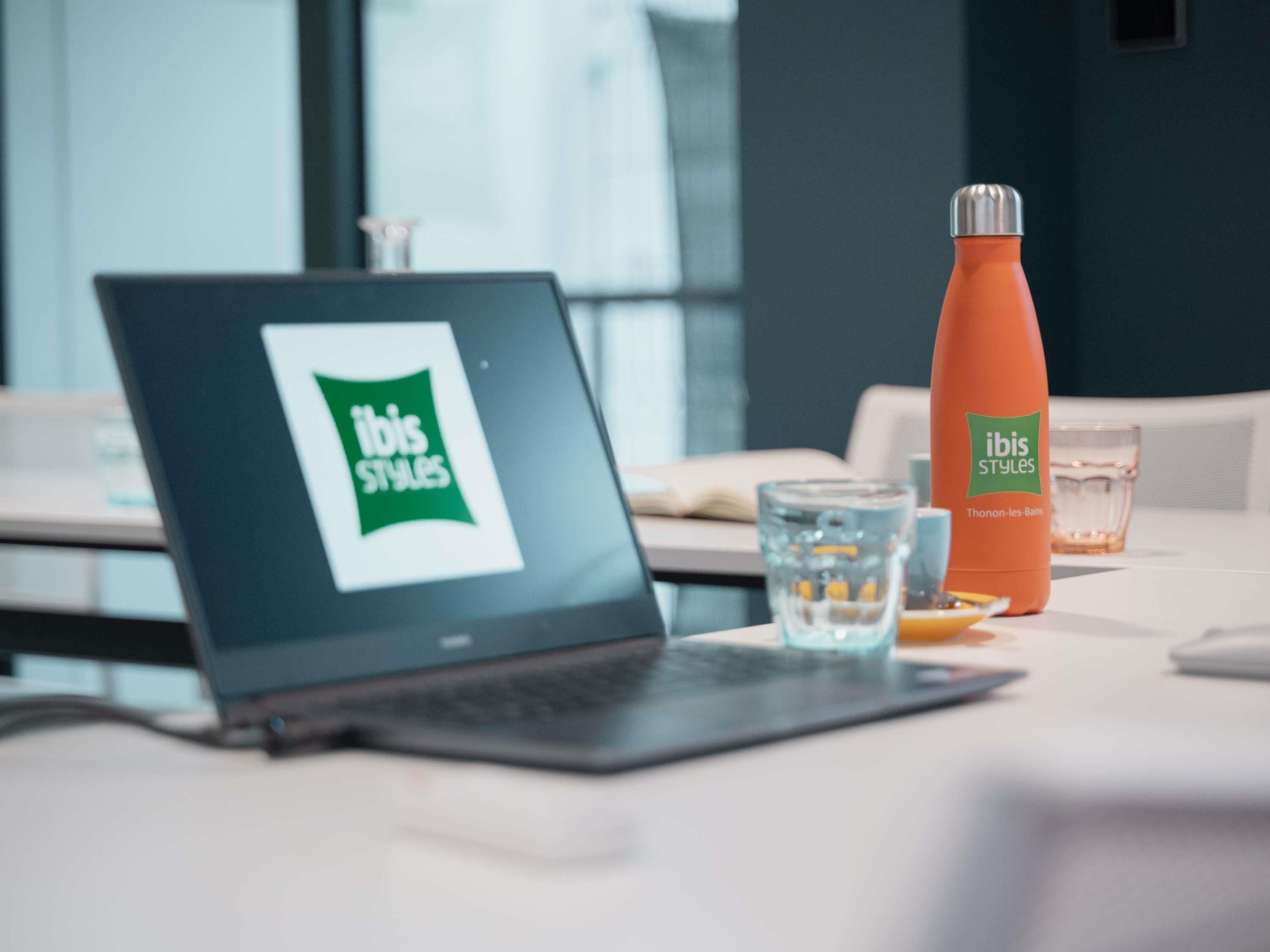 Ein Laptop mit Ibis Styles Logo, eine Wasserflasche und ein Glas auf einem Tisch.