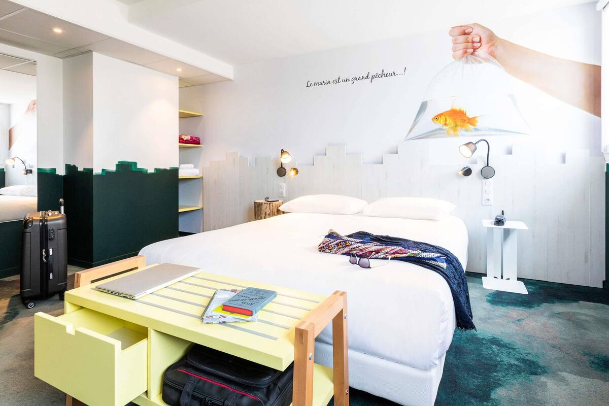 Quarto de hotel com uma cama, uma mesa com um laptop e livros, uma mala, uma luminária na parede e uma decoração com peixe.