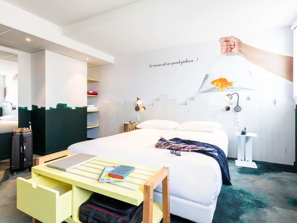 A hotel room with a bed, a desk with a laptop and books, a suitcase, a wall-mounted lamp, and a fish decoration. - ibis Styles Thonon Les Bains Hotel (Thonon-les-Bains)