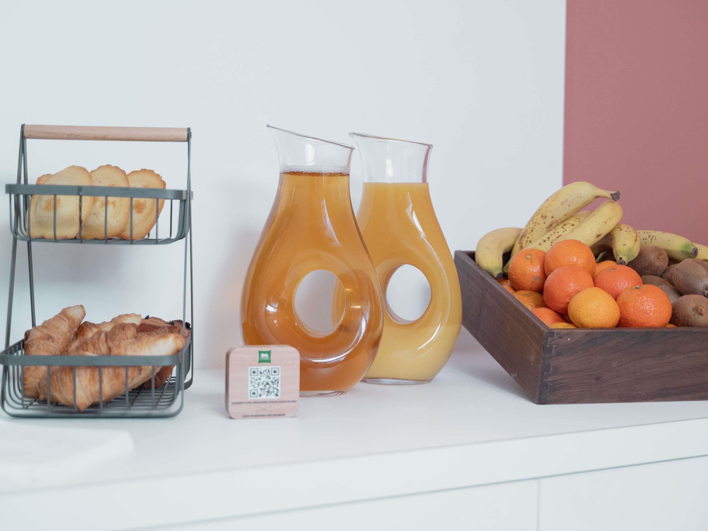Un comptoir de cuisine avec un présentoir à pain, deux carafes de jus d’orange, un code QR et un saladier rempli de bananes, d’oranges et de kiwis.