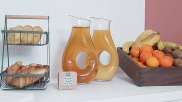 Un comptoir de cuisine avec un présentoir à pain, deux carafes de jus d’orange, un code QR et un saladier rempli de bananes, d’oranges et de kiwis.