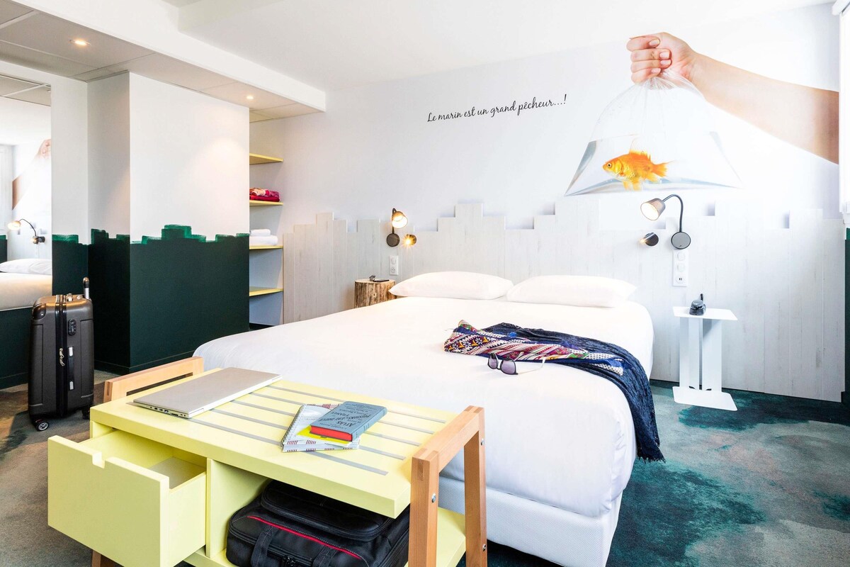 Ett hotellrum med en säng, ett skrivbord med en bärbar dator och böcker, en resväska, en vägglampa och en fiskformad dekoration.
