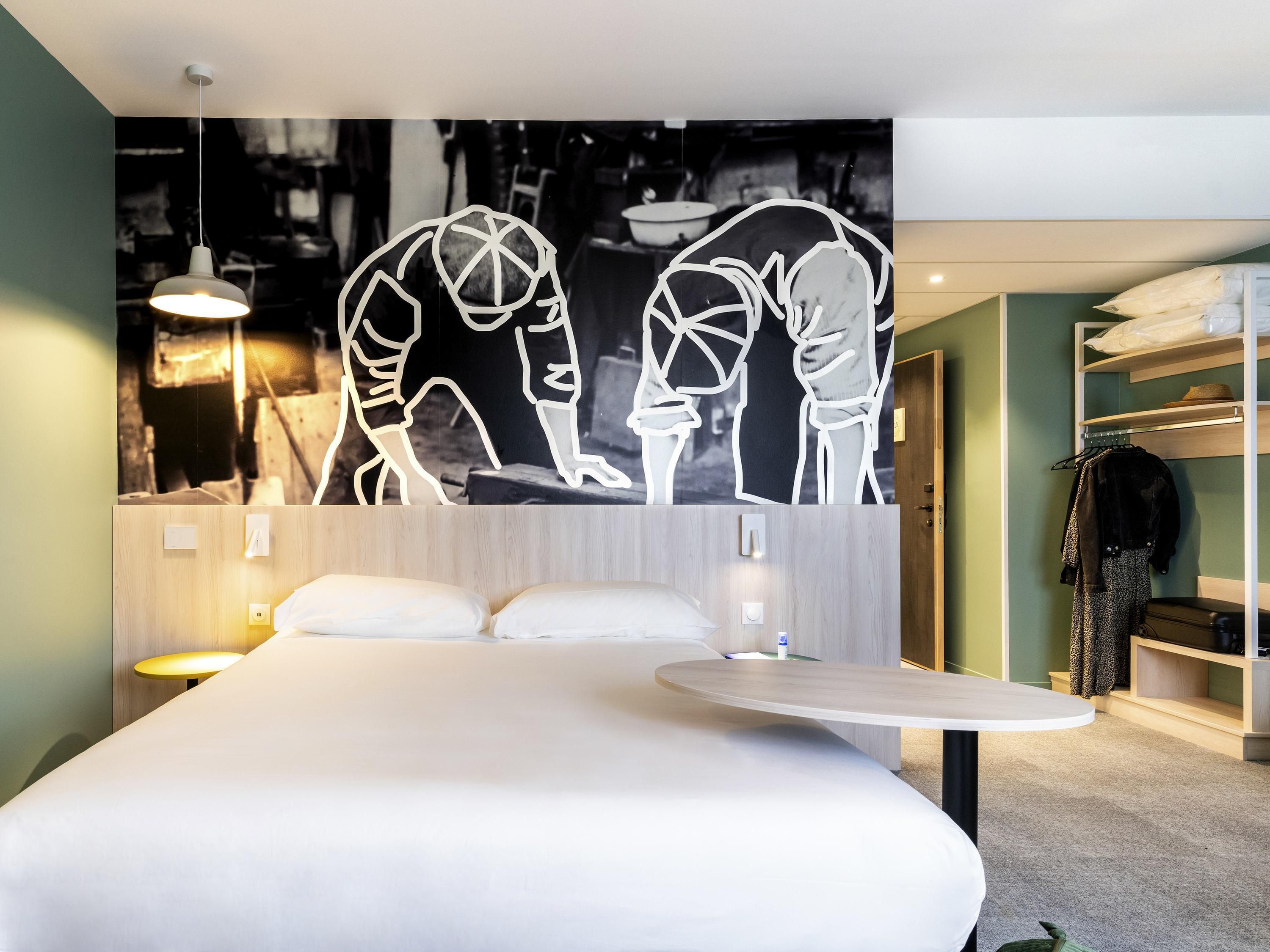A modern hotel room with a large bed, bedside tables, a round table, and a wall-mounted artwork.