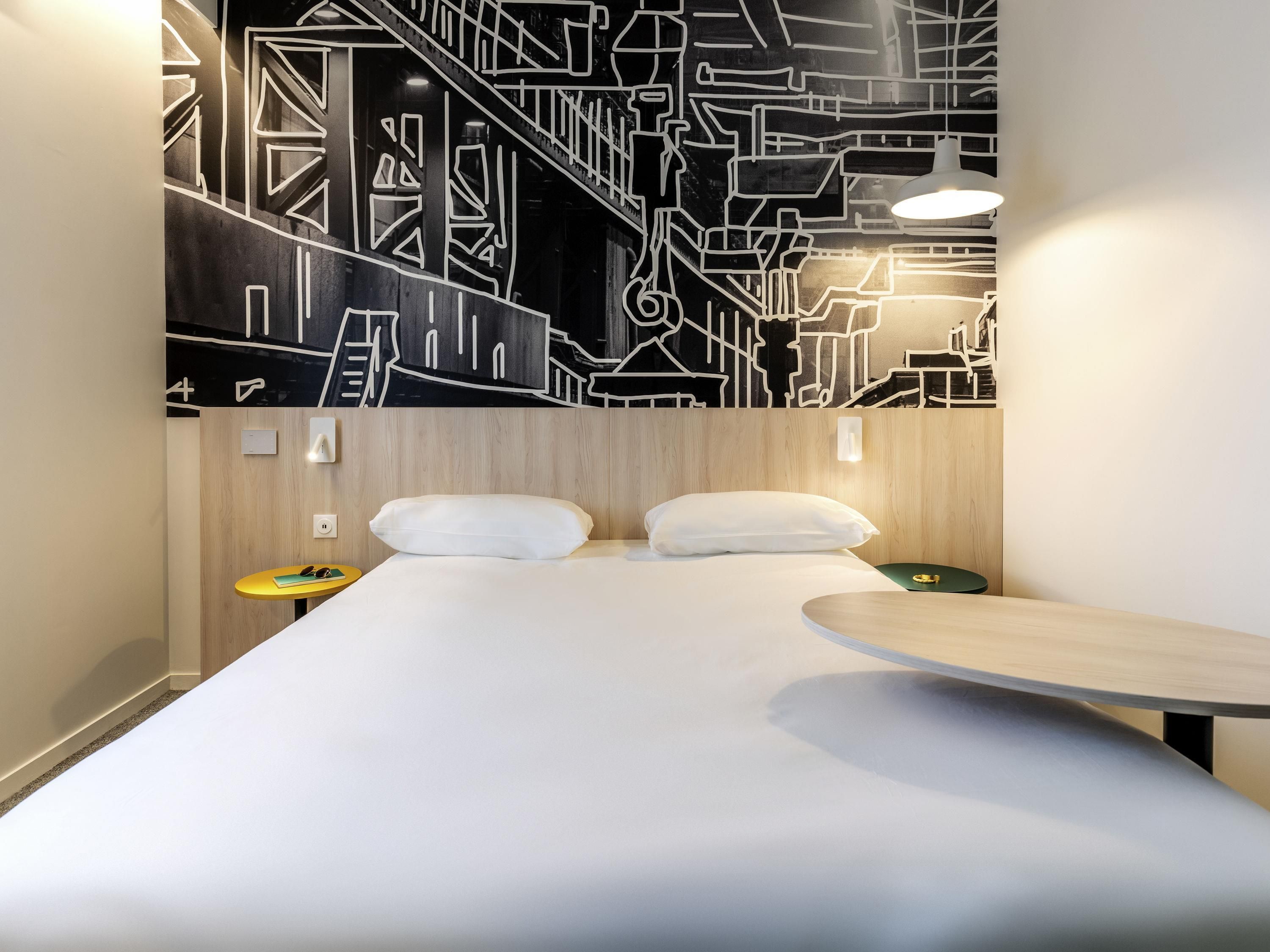 A modern hotel room with a bed, two pillows, a small table, a wall art piece, and a nightstand with a lamp.