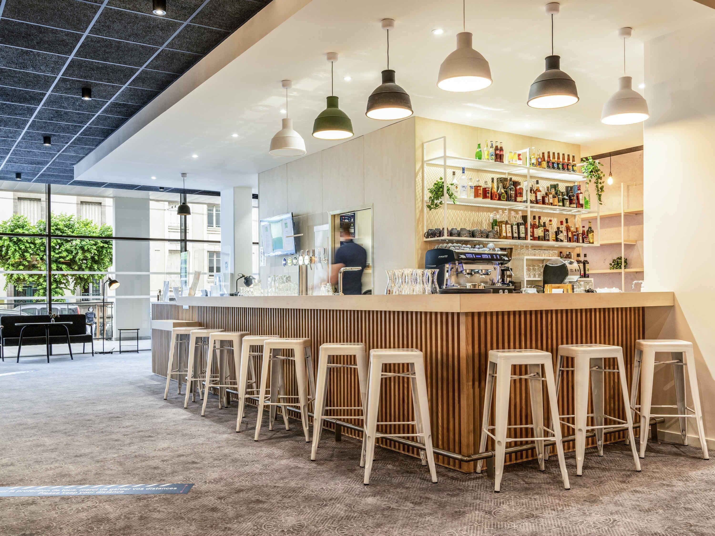 Uma área de bar moderna com bancada de madeira, banquetas brancas e iluminação pendente.