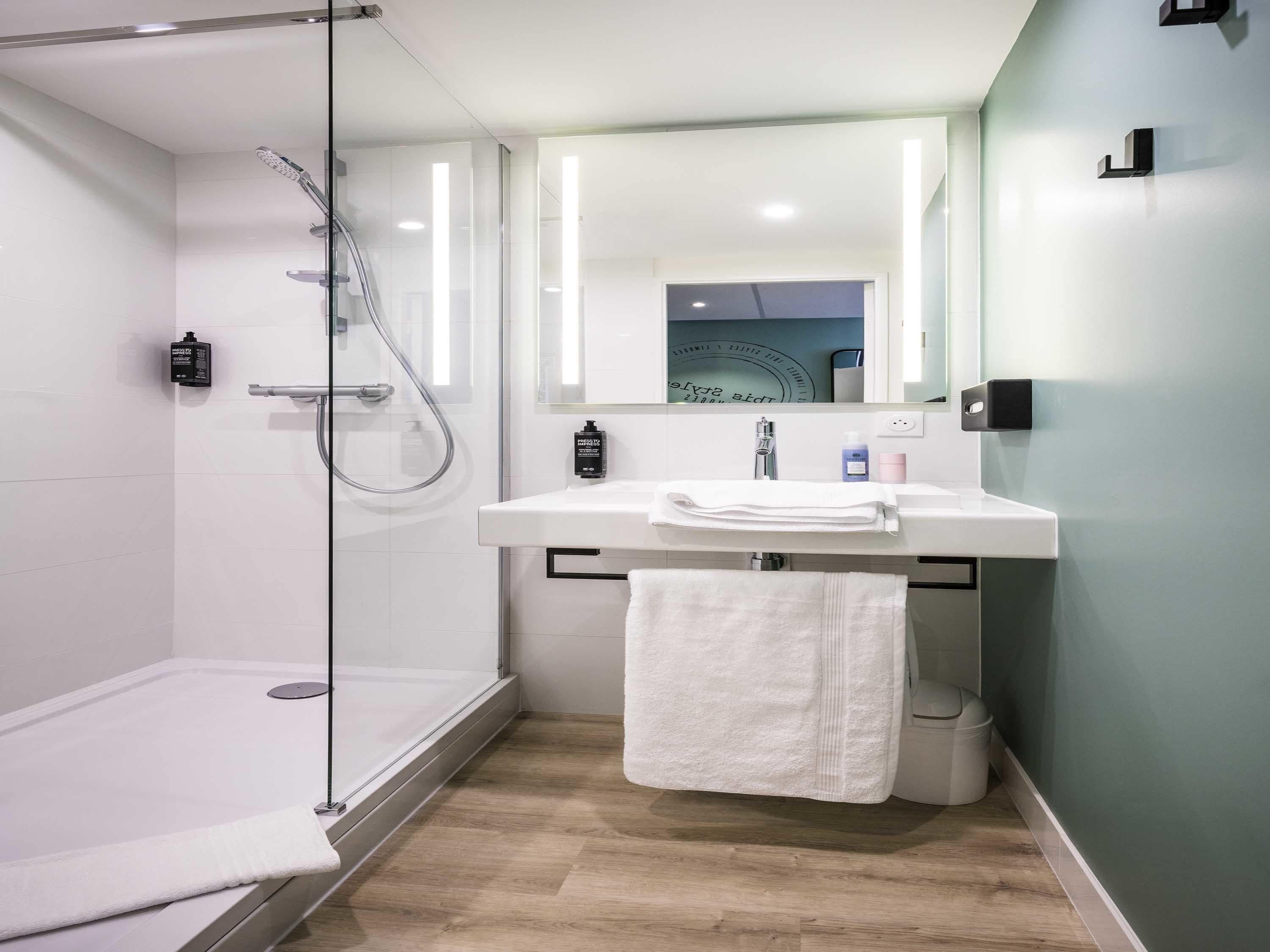 A modern bathroom with a glass shower enclosure, a white sink with a mirror above, and a wooden floor.