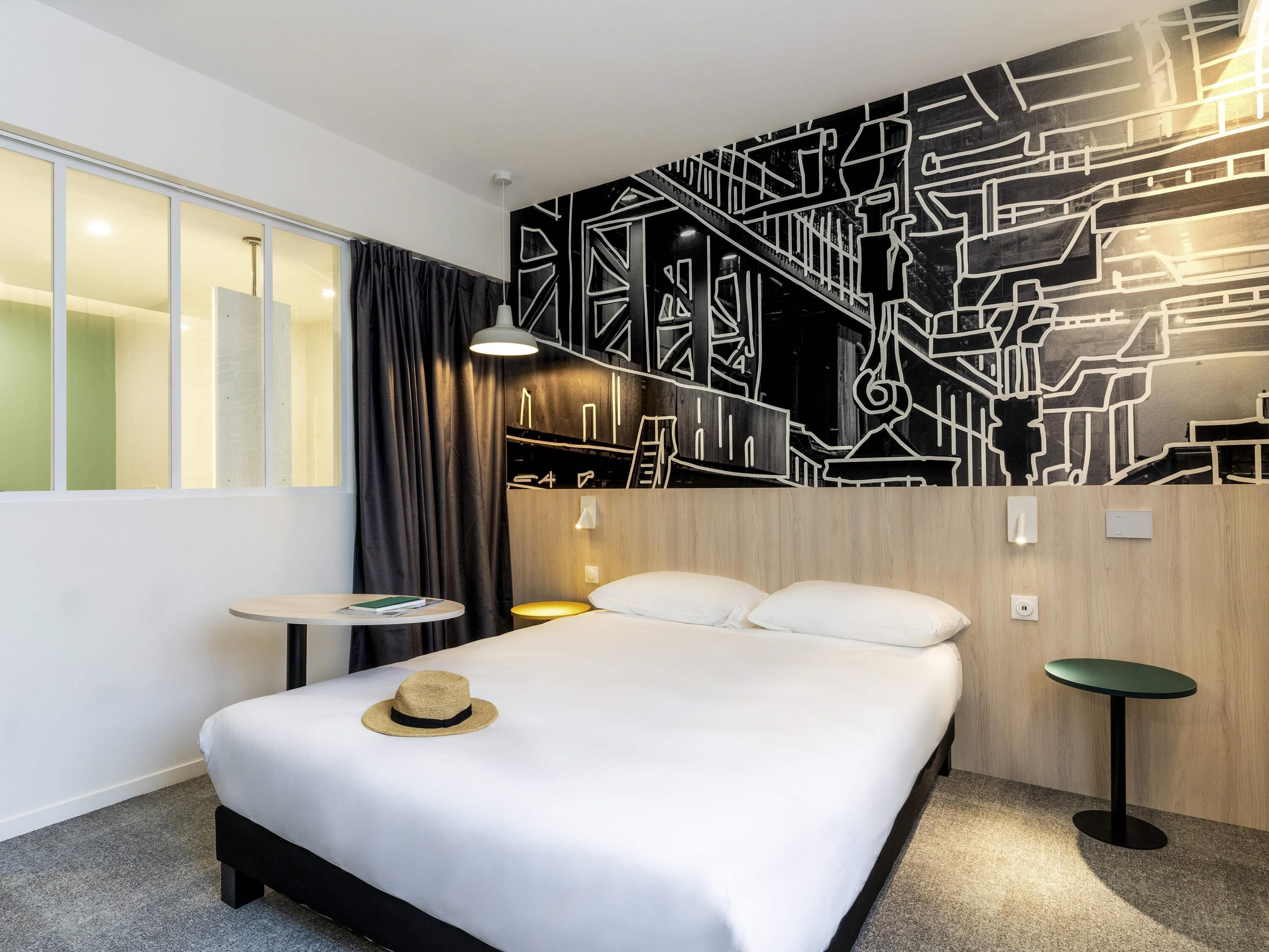 Una habitación de hotel moderna con una cama grande, una mesita, una silla y un mural en la pared.