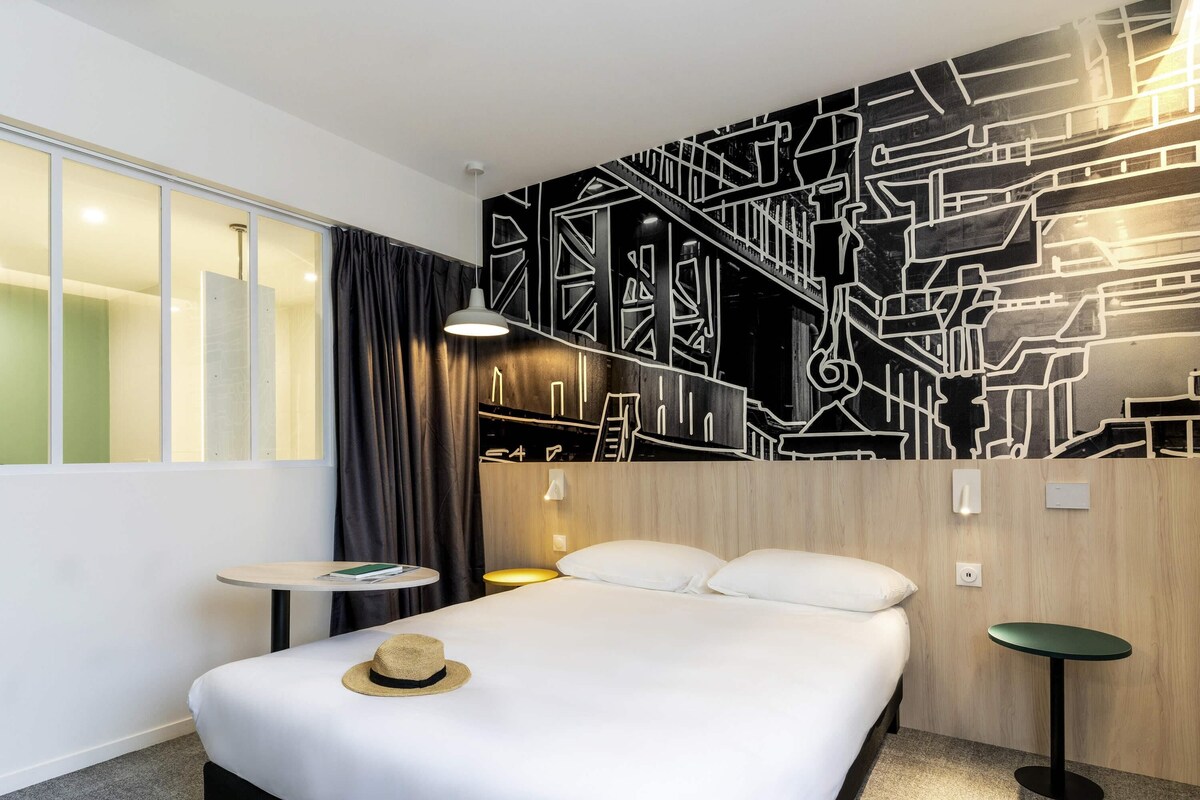 A modern hotel room with a large bed, a small table, a chair, and a wall mural.