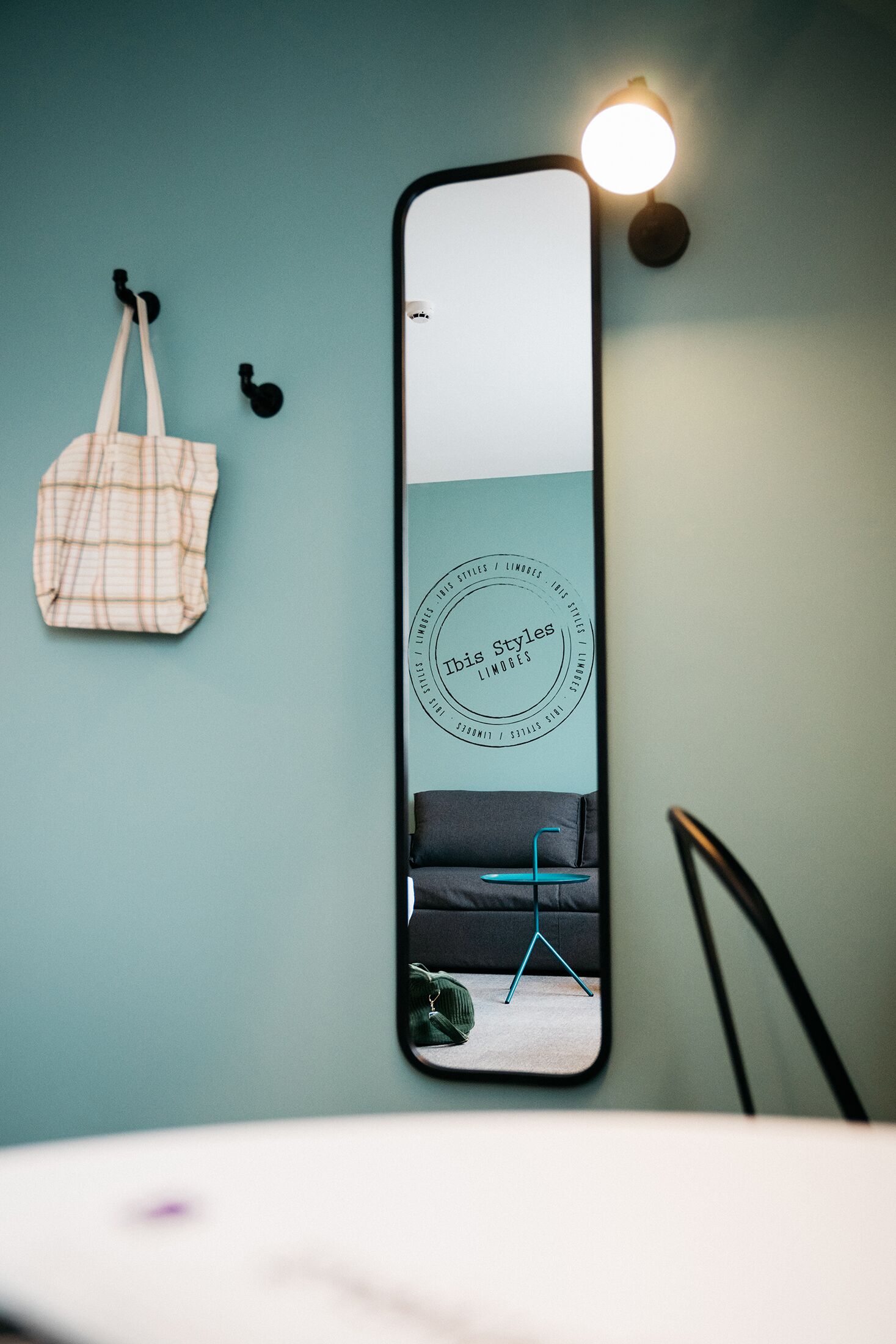 Um espelho reflete um quarto com uma parede verde-água, uma bolsa xadrez pendurada e uma cadeira preta.