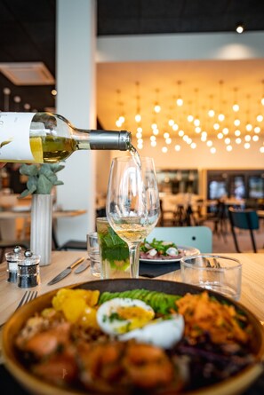 A dining table with a plate of food, a glass of white wine, and a bottle of wine being poured. - ibis Styles Limoges Centre (Limoges)