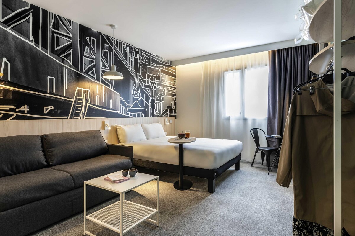 A hotel room with a bed, a sofa, a small table, a chair, a coat rack, and a large abstract wall mural.