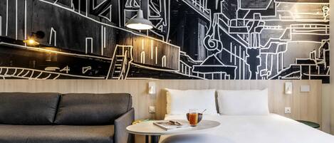 A modern hotel room with a large bed, a sofa, a small table with a drink, and a wall with a geometric pattern.