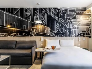 A modern hotel room with a large bed, a sofa, a small table with a drink, and a wall with a geometric pattern. - ibis Styles Limoges Centre (Limoges)