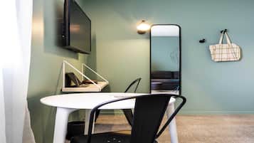Quarto pequeno e moderno com uma mesa branca, cadeira preta, TV fixada na parede, espelho na parede e uma bolsa pendurada.