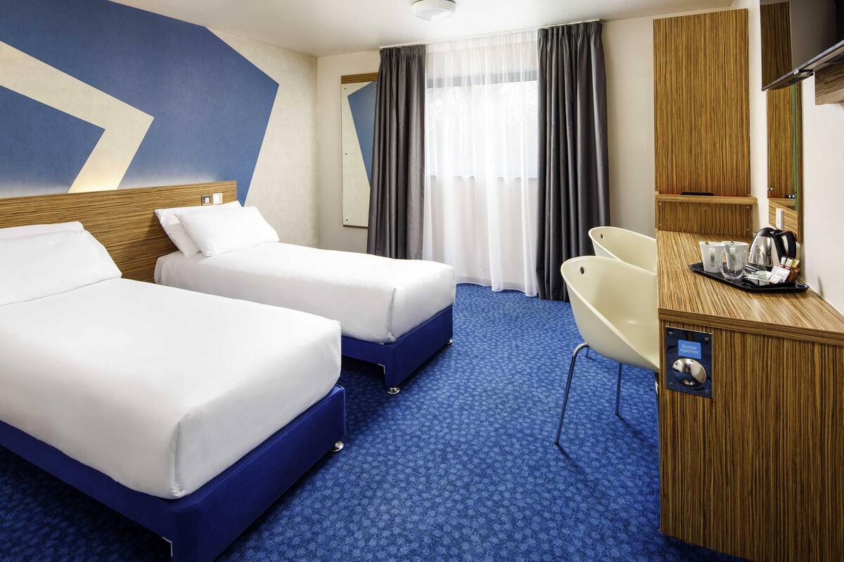 Ein Hotelzimmer mit zwei Betten, einem Schreibtisch mit Flaschenöffner, einem Sessel und einem großen Spiegel.
