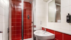 A bathroom with a glass-enclosed shower, a white sink, and a red-tiled wall. - Ibis Budget Heathrow Terminal 5 (Slough)