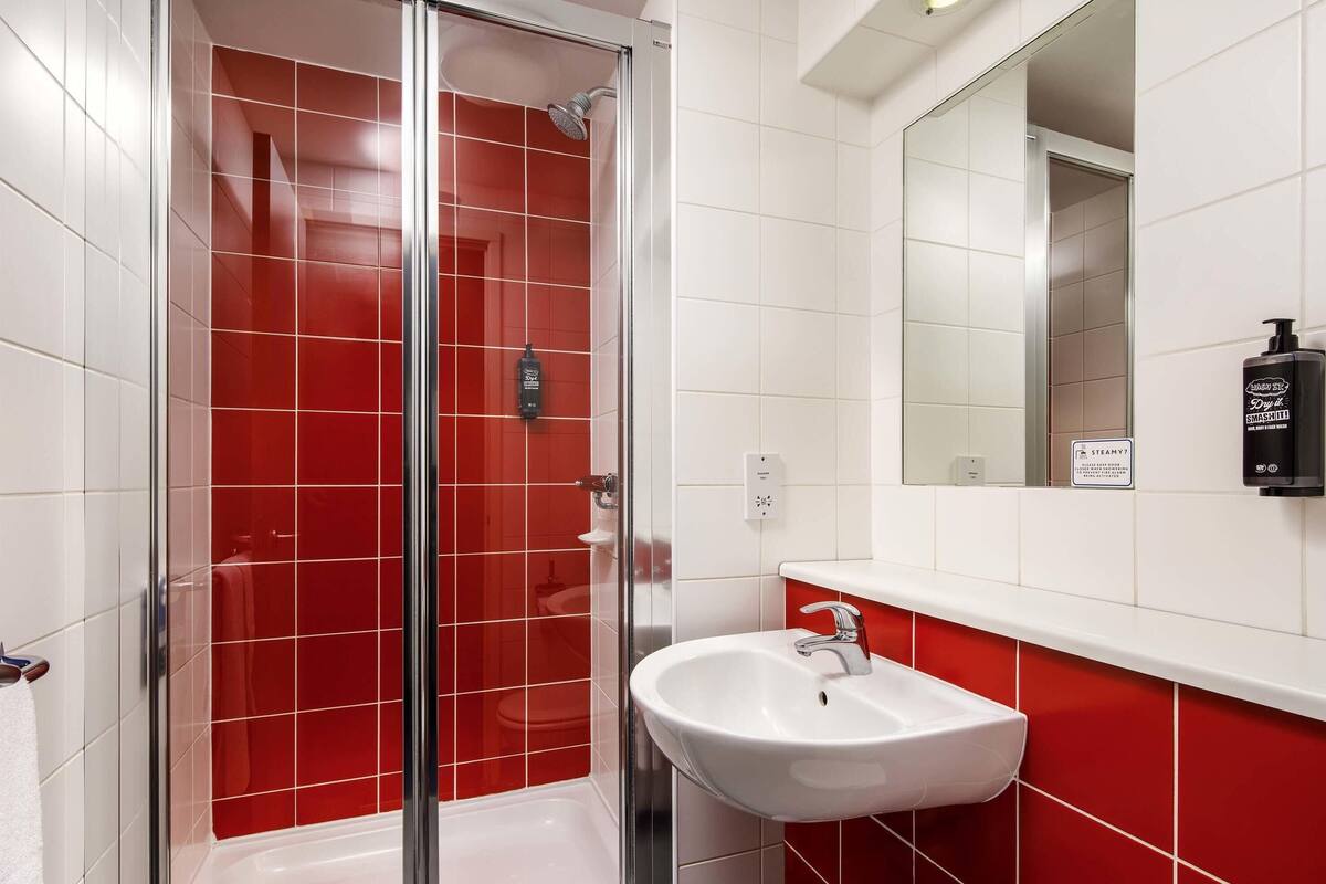 Ein Badezimmer mit einer gläsernen Duschkabine, einem weißen Waschbecken und einer roten Fliesenwand.