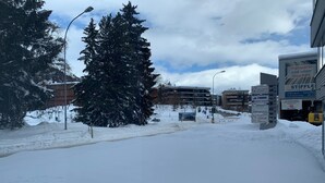 Una calle nevada en la ciudad con edificios, un gran árbol de Navidad y varios letreros de negocios.