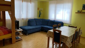 Una habitación con una cama litera, un sofá azul, una mesa de comedor con sillas y una ventana con persianas.