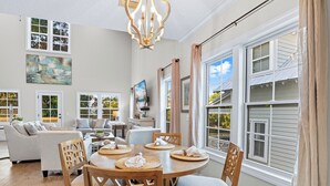 A spacious living room with a large wooden dining table, comfortable seating, and large windows offering views of the outside. - Spacious family fun with game room, only 4 miles from East Beach! Relax + Enoy! (St. Simons Island)