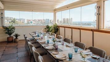 A dining area with a long table set for multiple guests, large windows offering a city view, and potted plants on the floor.