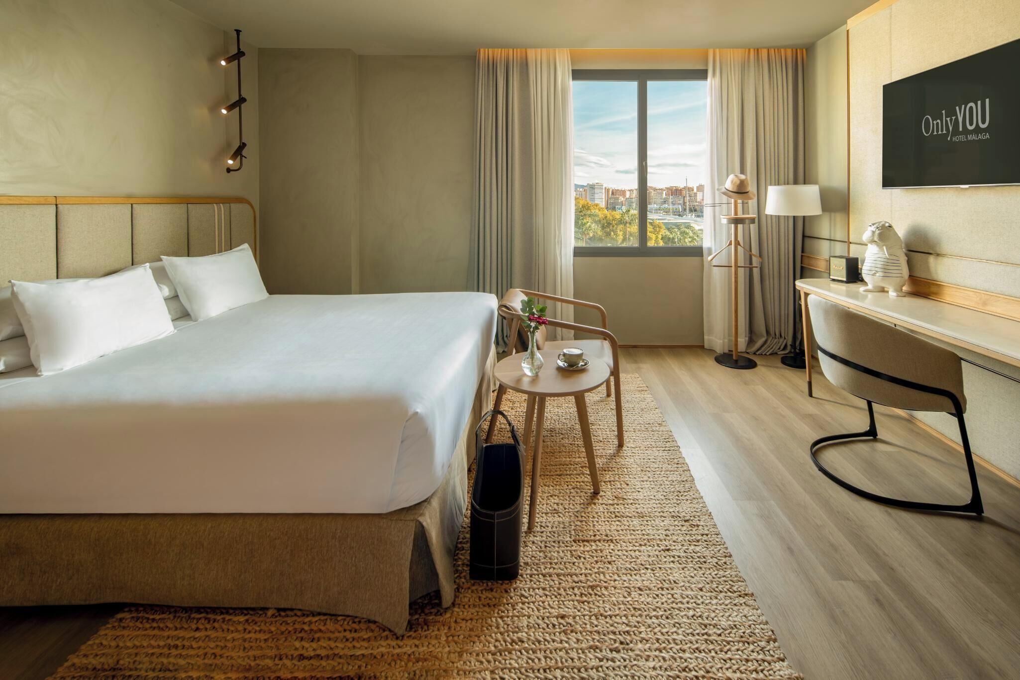 Habitación de hotel con una cama grande, un escritorio, una silla, una mesita con un jarrón y una taza, una lámpara y vistas a la ciudad desde la ventana.