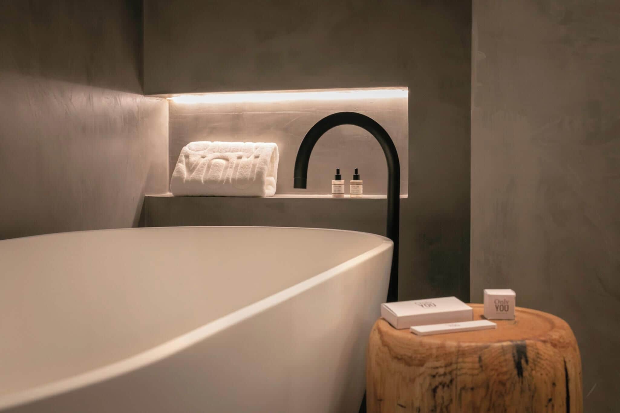 A modern bathroom with a large bathtub, a towel hanging on a wall-mounted rack, and toiletries on a wooden side table.