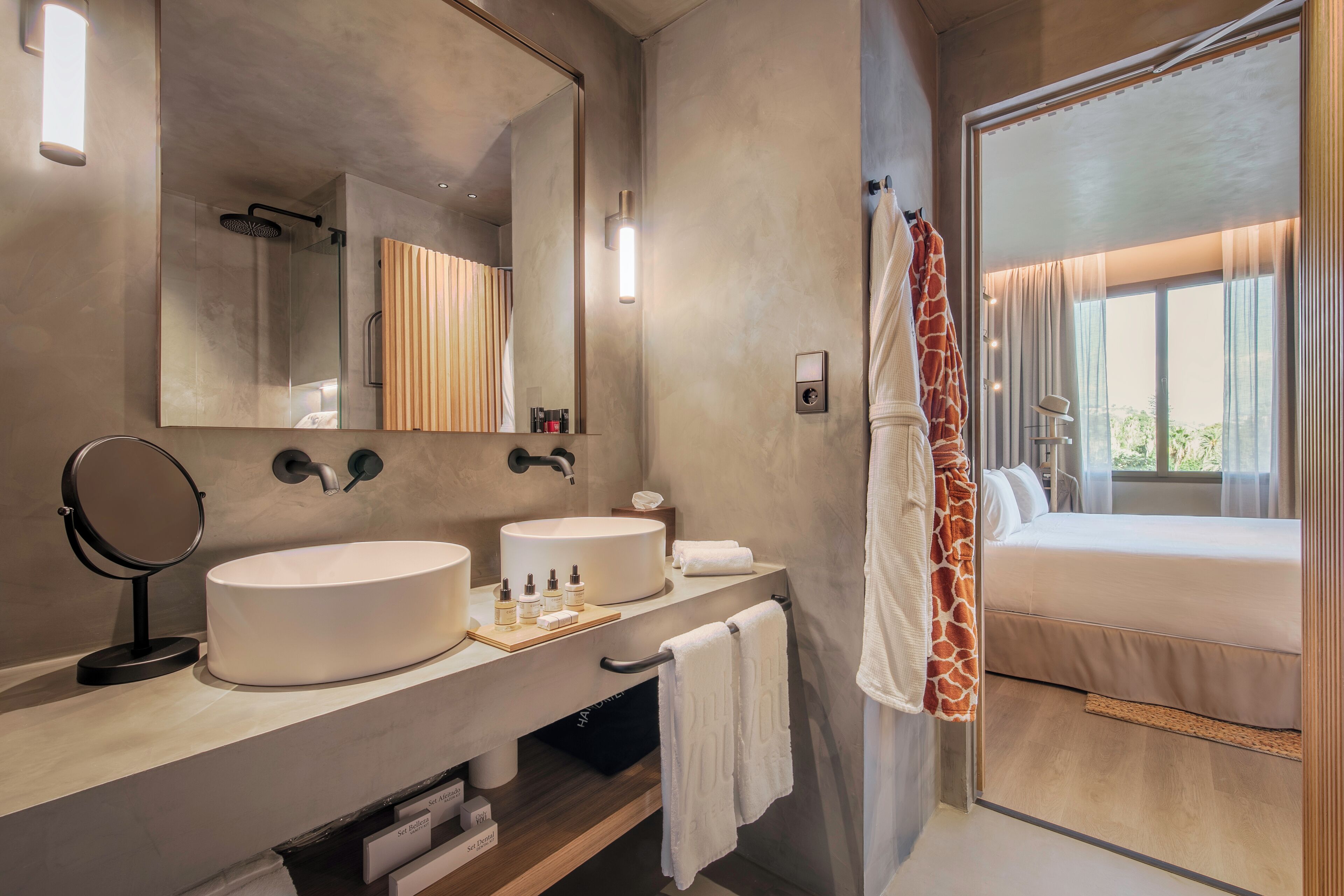 A modern bathroom with a large mirror, two sinks, a shower, and a bed with a view.