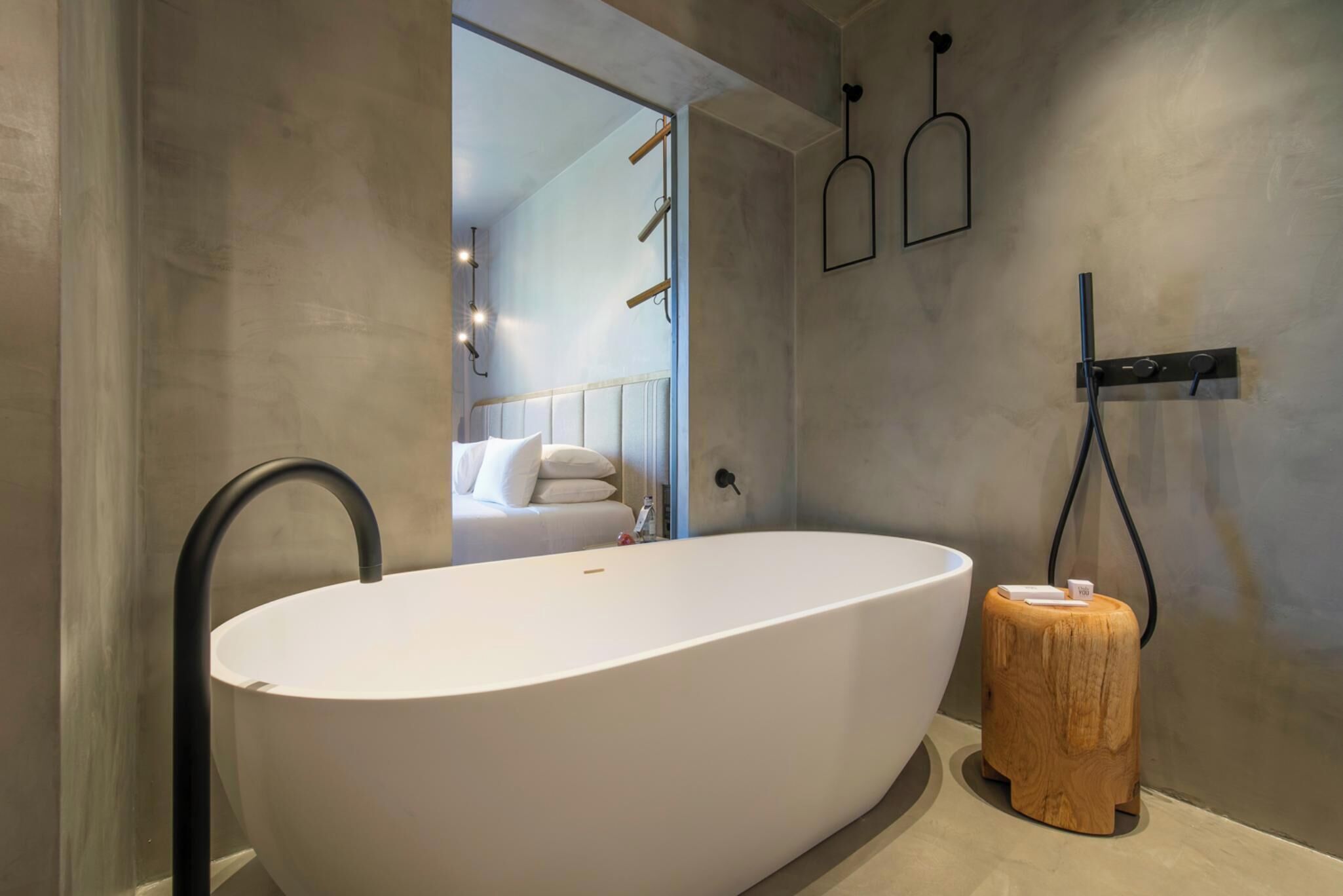 A modern bathroom with a freestanding bathtub, a wooden stool, and a mirror reflecting a bedroom.