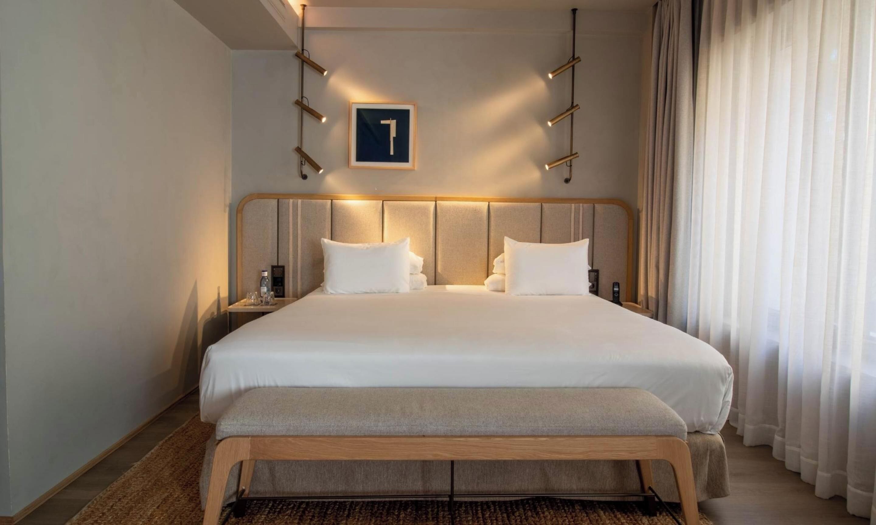 Habitación de hotel con una cama grande, una mesita de noche con una botella y vasos, un banco de madera, y un cartel con el número 1 en la pared.