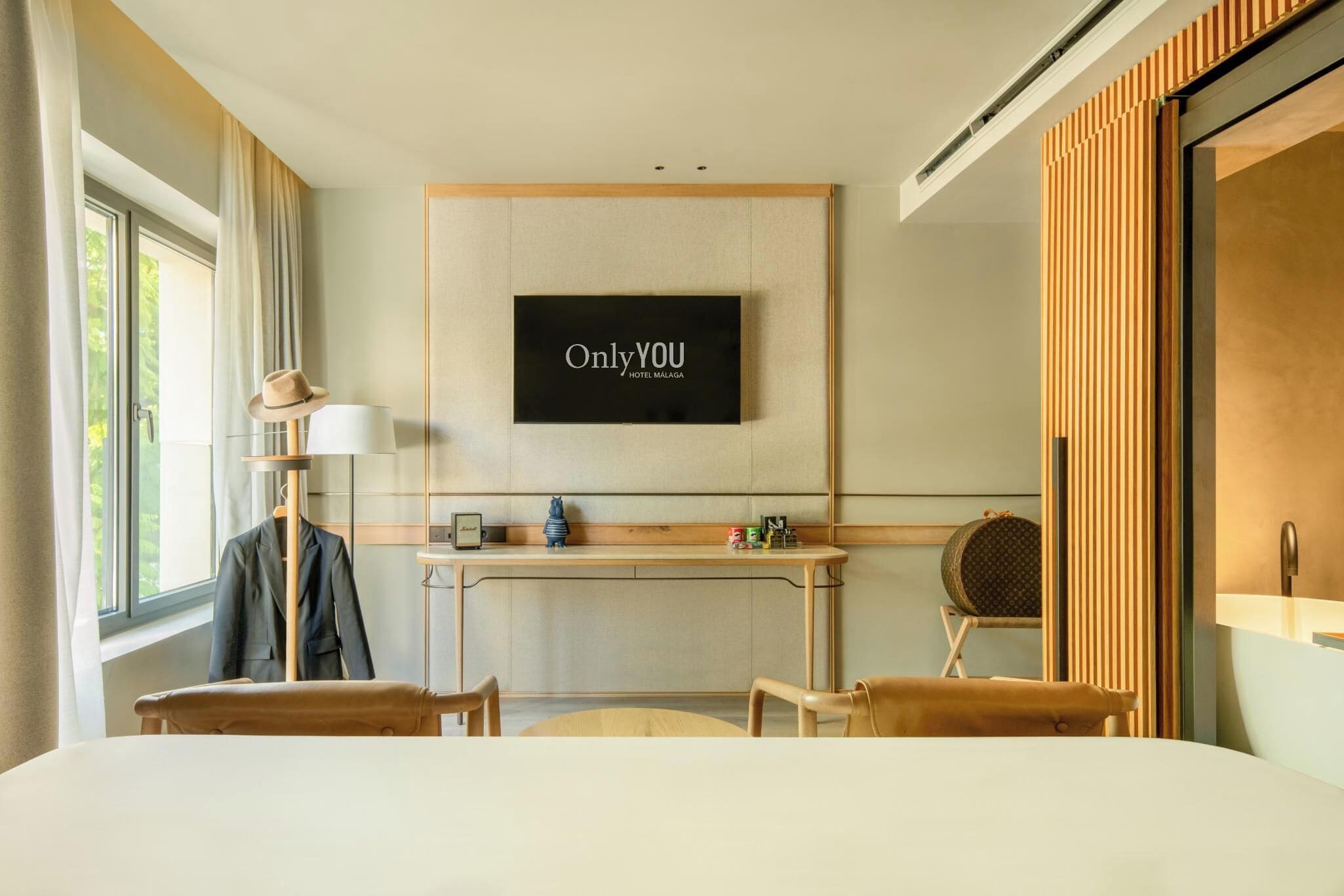 A hotel room with a bed, a desk, a chair, a television mounted on the wall, and a window with curtains.