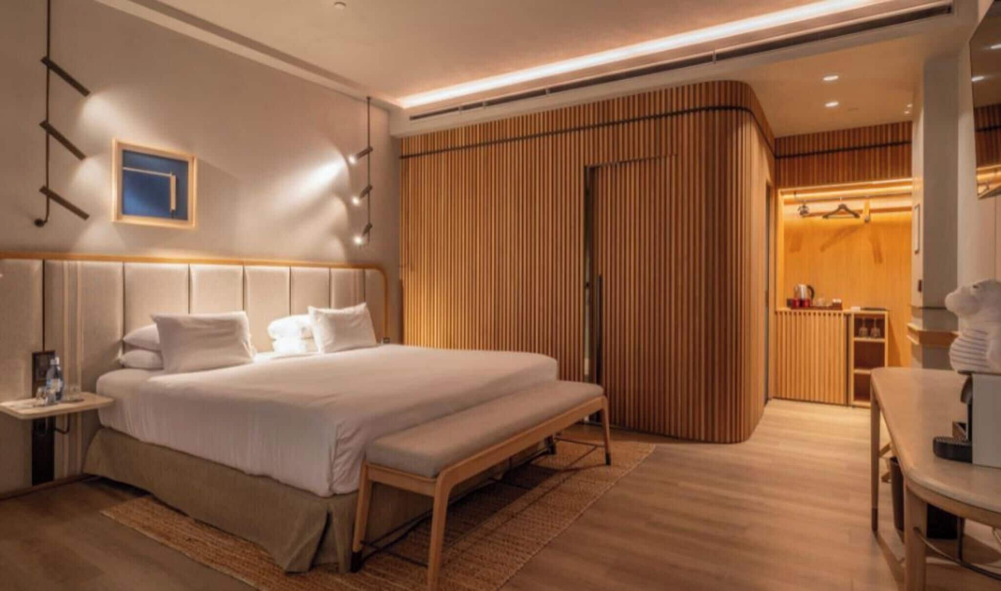 Quarto de hotel moderno com uma cama grande, mesas de cabeceira, um banco, um guarda-roupa e uma pequena área de estar.