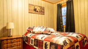 Un dormitorio con pared de madera, una cama con colcha estampada con motivos de animales, una mesita de noche con lámpara y una ventana con cortinas.