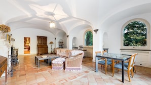 A spacious living room with a high ceiling, a dining area with a glass table, and a view of greenery through large windows. - Deluxe Triple Library Room (SORRENTO)