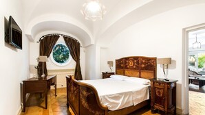 A bedroom with a wooden bed, a desk, a chair, a window with curtains, and a chandelier. - Deluxe Triple Library Room (SORRENTO)