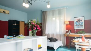 A room with a bed, a desk, a chair, a small table, a shelf with books, a vase with flowers, and a poster on the wall.