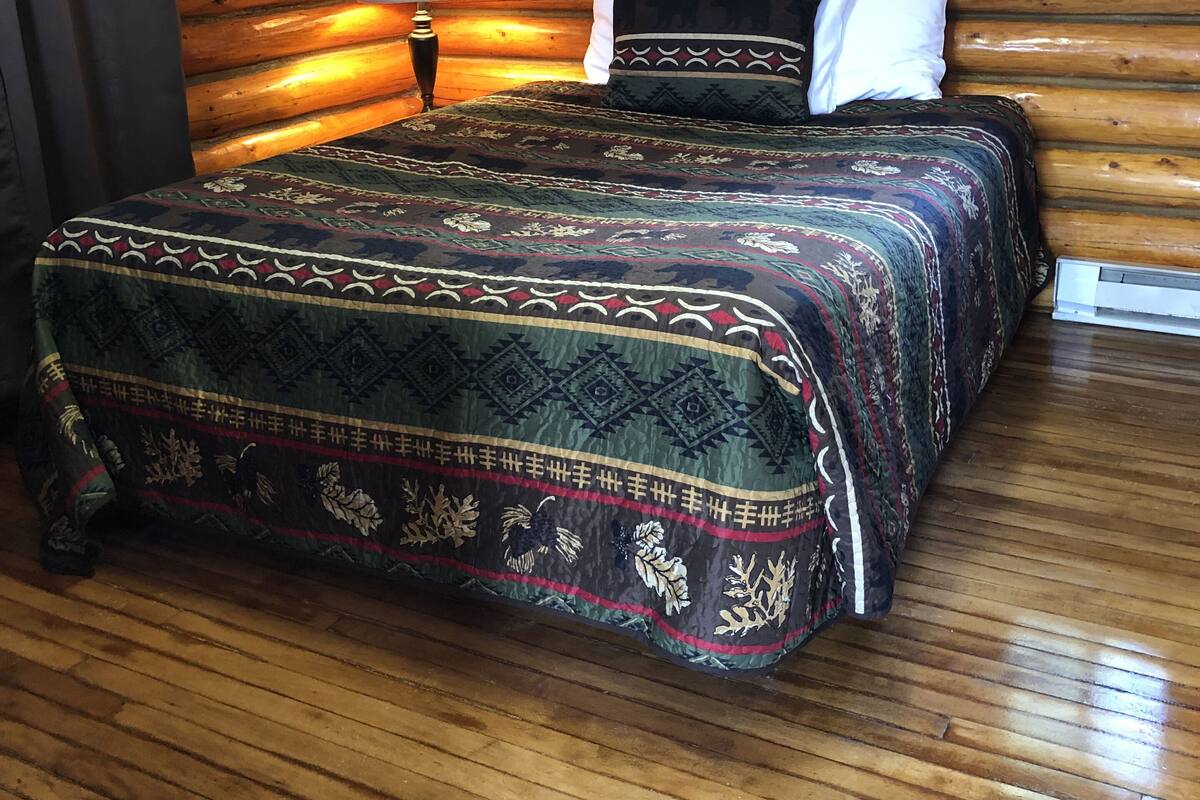A log cabin-style room with a wooden floor, a bed with patterned bedding, a lamp, and a window with red curtains.