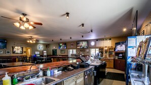 A bar area with a counter, sinks, and a ceiling fan. There are tables and chairs in the background. - Sportsmans Lodge (Ennis)