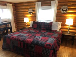 Quarto de cabana de tronco com cama, duas mesas de cabeceira com abajures, uma escrivaninha e uma cadeira.