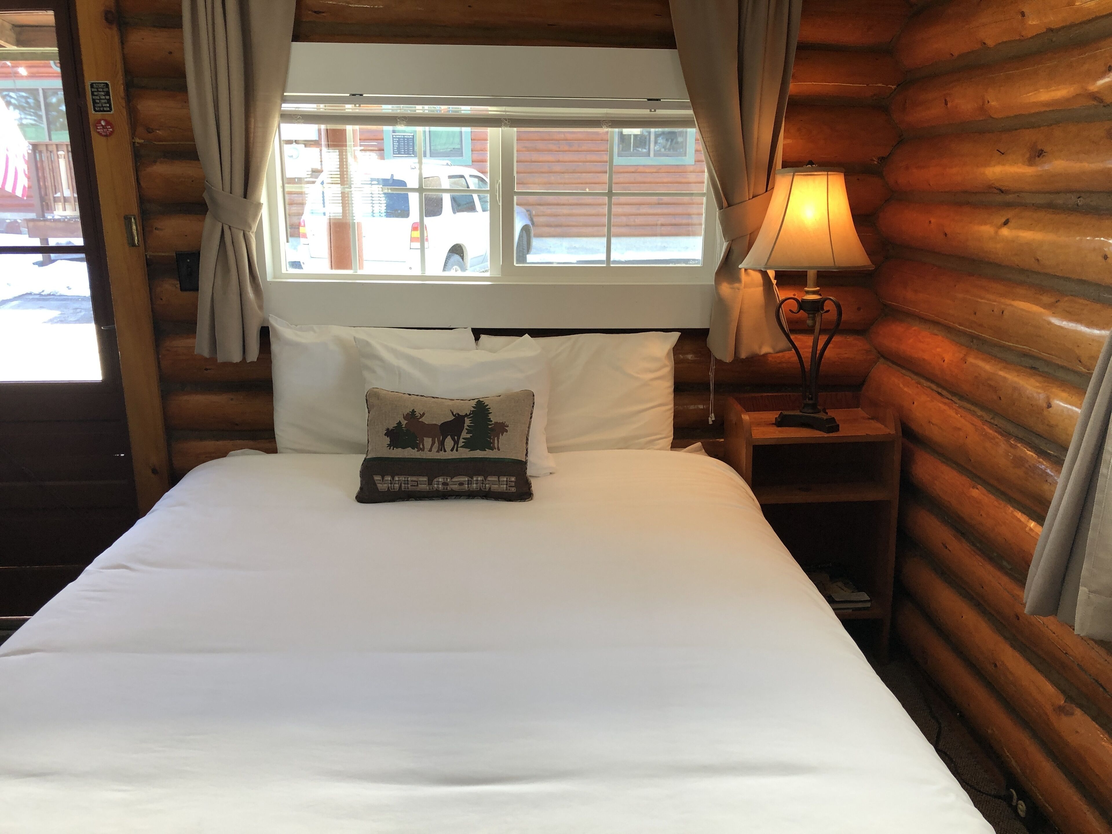 Une chambre de style rustique avec un lit, une fenêtre avec des rideaux, une lampe sur une table de chevet et un oreiller orné d’un message de bienvenue.