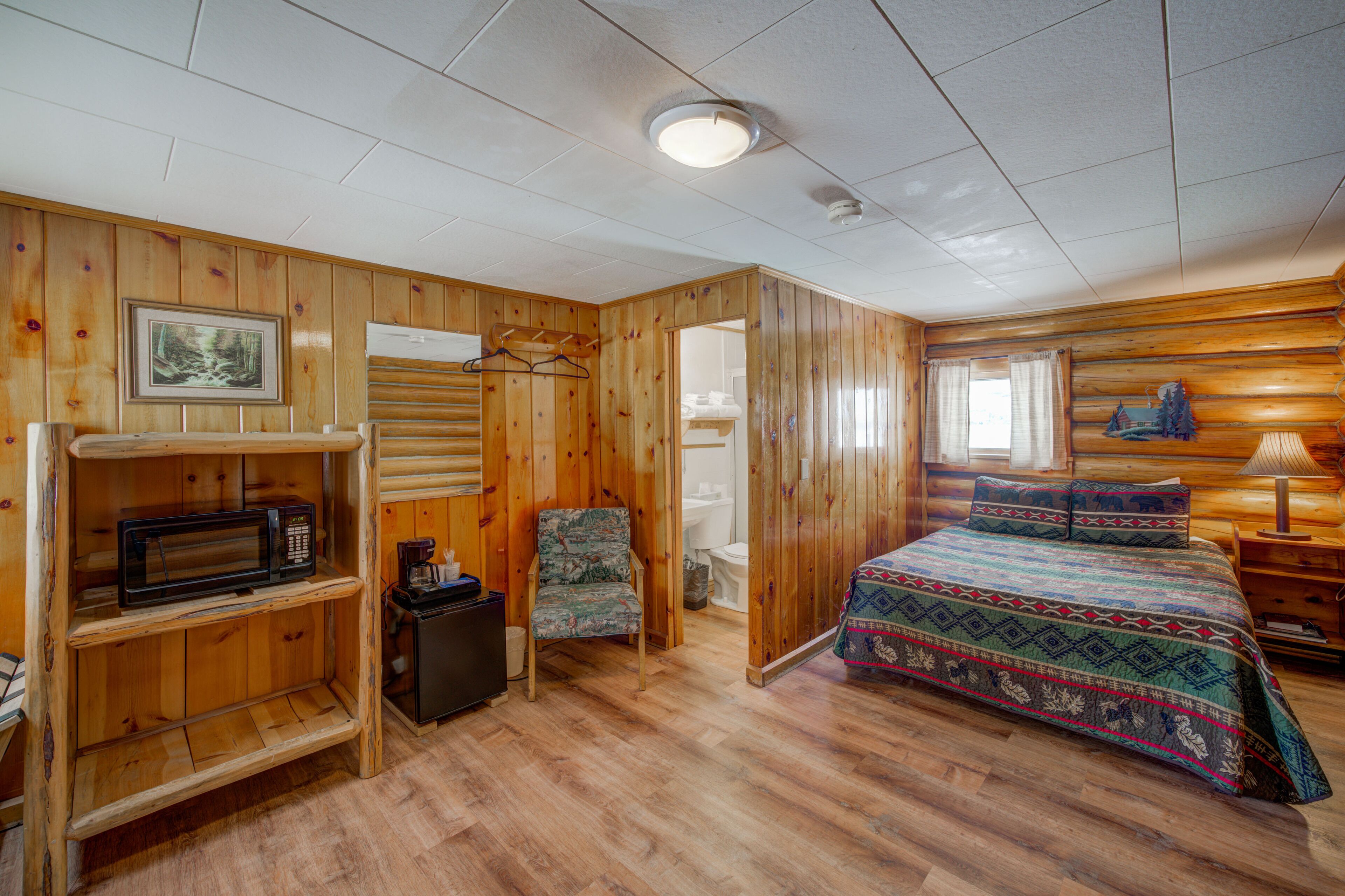 A wooden cabin room with a bed, a chair, a shelf with a microwave, and a picture on the wall.